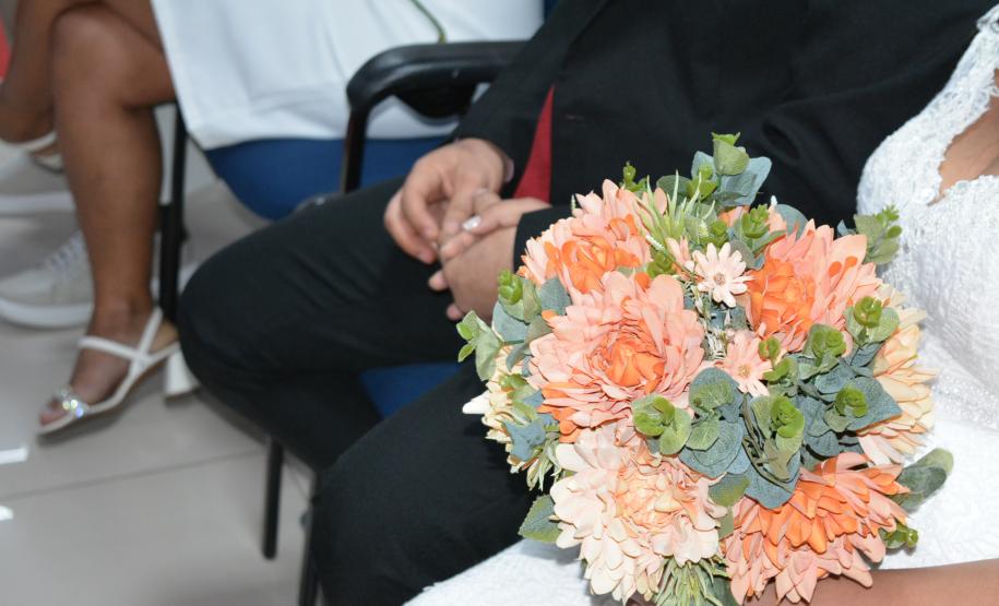 Close nas mãos de uma noiva segurando um buquê de flores em tons de pêssego, alaranjado e folhagens verdes. Ela veste um vestido branco com detalhes em renda e está sentada ao lado de um homem de terno preto e gravata vermelha.