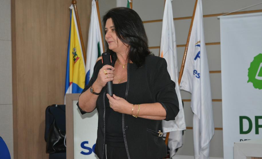 Uma mulher de cabelos escuros e conjunto preto segura um microfone enquanto fala. Logo atrás dela, estão posicionadas mastros com bandeiras oficiais e parte de um banner da Defensoria Pública.