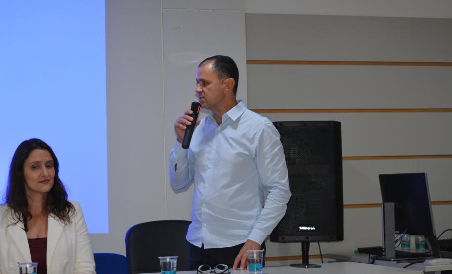 Um homem de camisa azul clara fala ao microfone em pé. Ao lado, uma mulher de blazer branco está sentada à mesa. Ao fundo, há uma tela de projeção exibindo uma luz azul e uma caixa de som preta.