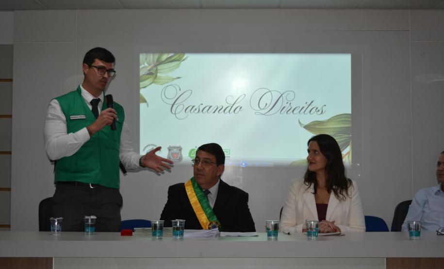 Um homem de colete verde fala ao microfone em pé ao lado de uma mesa com três pessoas sentadas. Atrás deles, uma projeção exibe a frase "Casando Direitos". Na mesa, destaca-se um homem com uma faixa verde e amarela no peito.