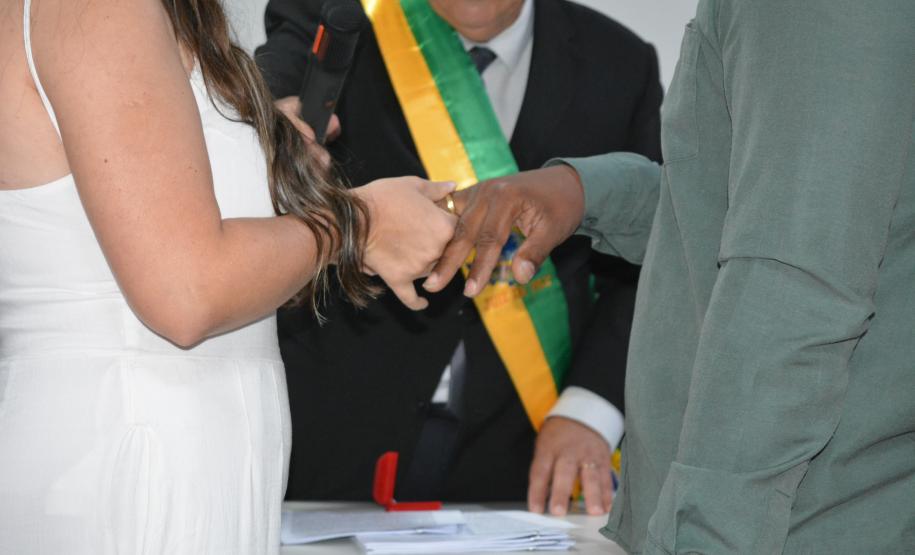 Close de um casal trocando alianças em uma cerimônia. A noiva veste branco e o noivo veste uma camisa verde. Ao fundo, o celebrante que segura o microfone usa uma faixa verde e amarela, e uma caixinha vermelha de anel repousa sobre a mesa.