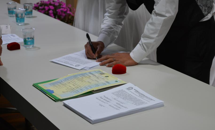 Foco nas mãos de uma pessoa de camisa branca e colete escuro assinando documentos sobre uma mesa clara. Próximo às mãos, há certidões de casamento com brasão oficial, caixinhas vermelhas para alianças e copos de água.