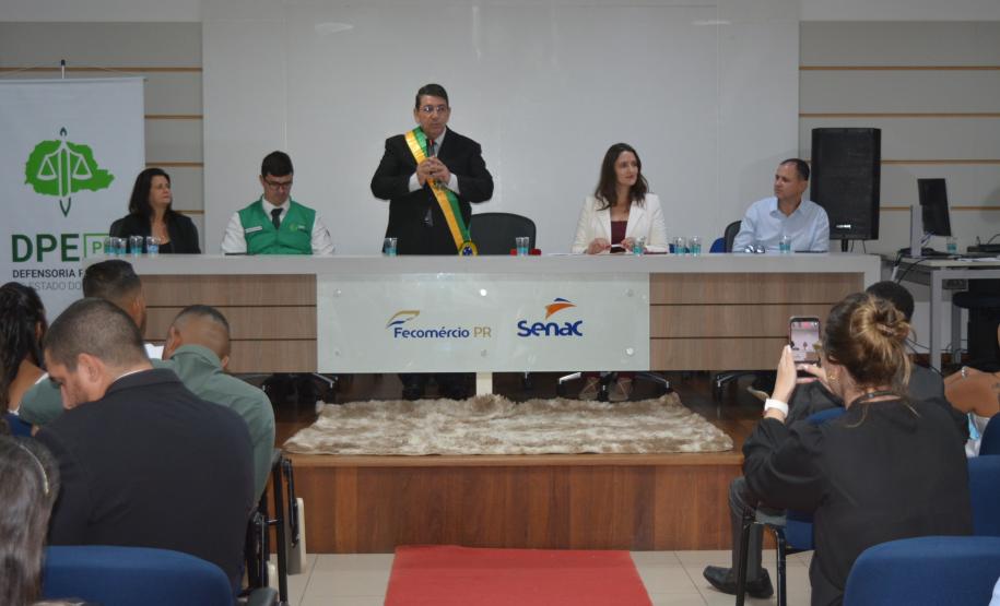 Visão ampla de um auditório onde um celebrante com faixa verde e amarela fala ao microfone em uma mesa diretiva com as logos do Fecomércio PR e Senac. À esquerda, há um banner da DPE-PR. O público assiste sentado, com pessoas fotografando a cena.