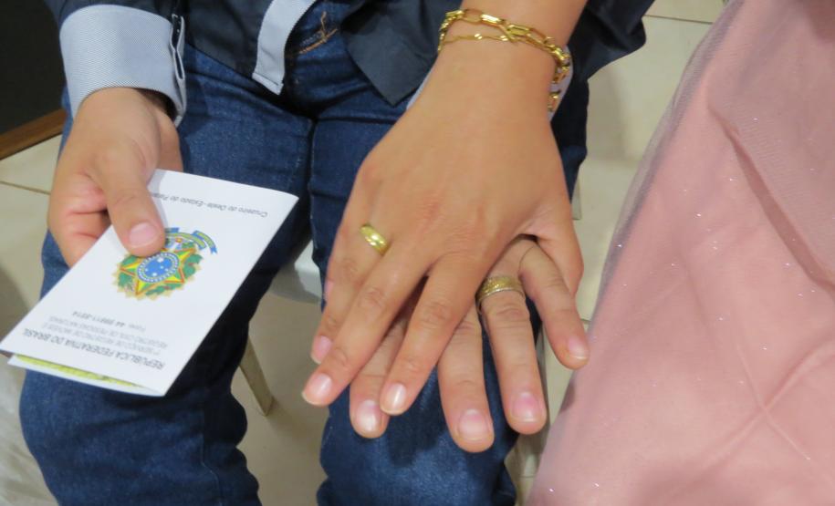 Foco nas mãos cruzadas de um casal em que ambos exibem alianças douradas. Uma das pessoas veste calça jeans e segura um documento da "República Federativa do Brasil", enquanto parte de um vestido rosa brilhante aparece à direita.