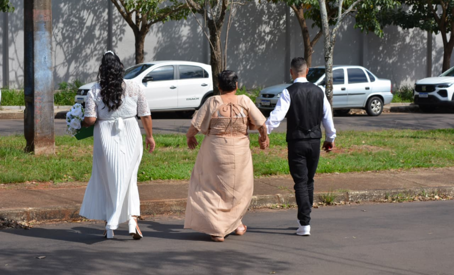Visão de costas de três pessoas caminhando de mãos dadas por uma rua asfaltada e arborizada com carros estacionados. Da esquerda para a direita, caminham uma noiva de vestido branco, uma mulher de vestido longo bege e um noivo de colete preto e camisa branca.