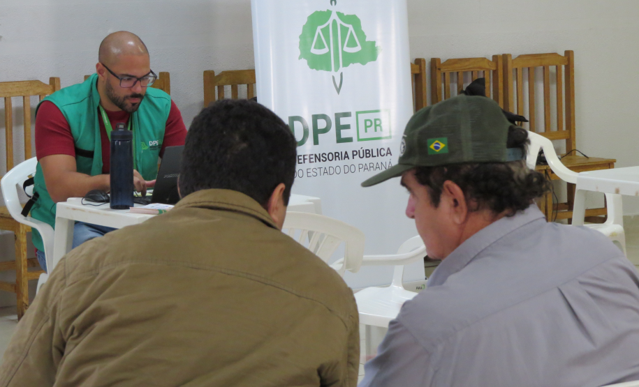 Um servidor de óculos e colete verde atende em um notebook numa mesa branca. À sua frente, dois homens aguardam sentados de costas. Ao fundo, há um banner vertical da DPE-PR.
