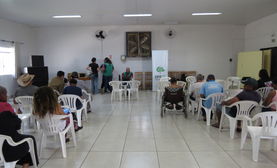 Visão ampla de um salão com diversas pessoas sentadas em cadeiras brancas voltadas para o fundo. Lá, servidores da DPE-PR estão próximos a mesas posicionadas ao lado de um banner da instituição.