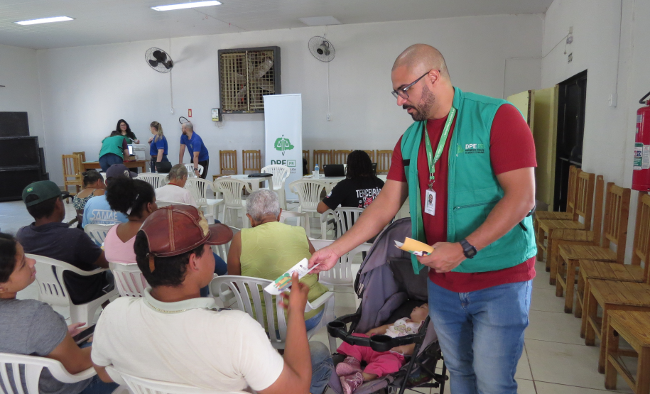Um funcionário em pé, de óculos e colete verde, entrega um panfleto a um homem sentado de costas. Ao lado do servidor há um carrinho com um bebê dormindo, e outras pessoas aguardam ao fundo do salão.