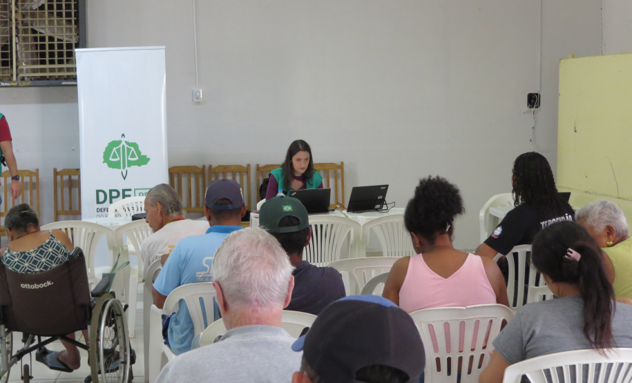 Uma mulher de colete verde trabalha em um notebook ao fundo da sala, ao lado de um banner da DPE-PR. À frente dela, diversas pessoas aguardam sentadas de costas, incluindo uma pessoa em cadeira de rodas.