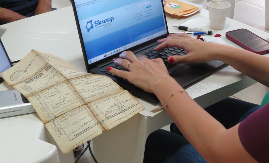 Fotografia em close de mãos femininas com esmalte vermelho digitando em um notebook que exibe a página do sistema Sinesp. Sobre a mesa branca, à esquerda do teclado, destaca-se um documento oficial muito antigo, amarelado e com marcas de dobras. Canetas, um celular e um copo descartável também compõem a cena.