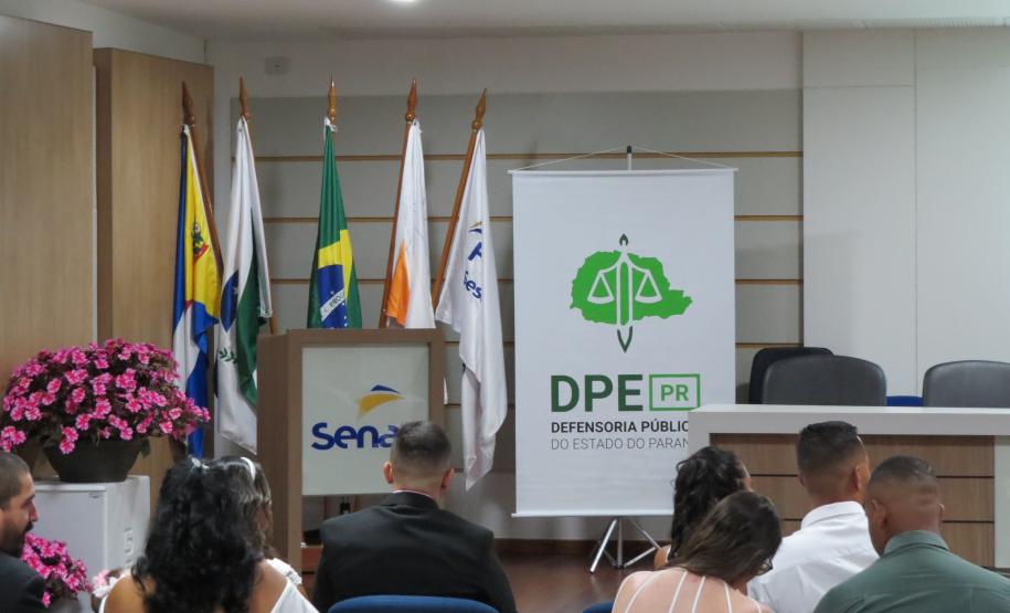 Um auditório com o público sentado de costas em cadeiras azuis, voltado para um palco. No palco, há um púlpito com a logo do Senac acompanhado por bandeiras oficiais, e à direita destaca-se um banner da Defensoria Pública (DPE-PR).