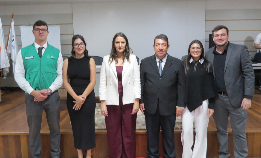 Seis pessoas posam lado a lado em um palco de piso de madeira com um tapete vermelho no centro. O grupo é formado por homens e mulheres em trajes sociais, incluindo, à extrema esquerda, um homem vestindo o colete verde da DPE-PR.