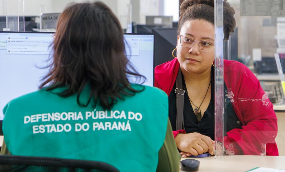 Imagem mostra servidor de costas com colete verde onde se lê Defensoria Pública do Parané e ao fundo uma mulher assistida durante Mutirão Meu Pai Tem Nome.