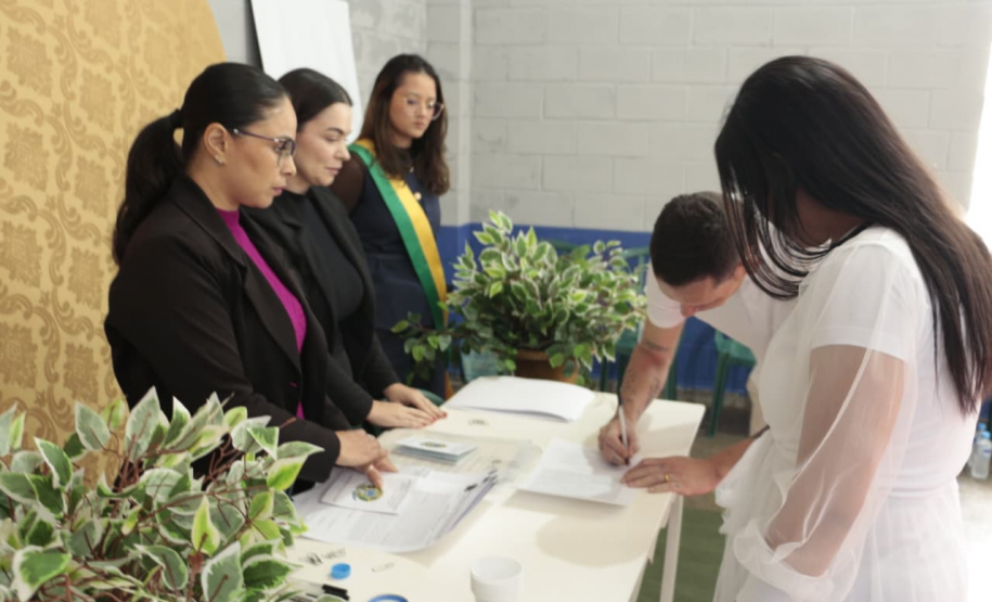 Um homem de camiseta branca assina um documento sobre uma mesa, ao lado de uma mulher de vestido branco. Atrás da mesa, três mulheres observam atentamente, sendo que uma delas usa uma faixa transversal com as cores da bandeira do Brasil.