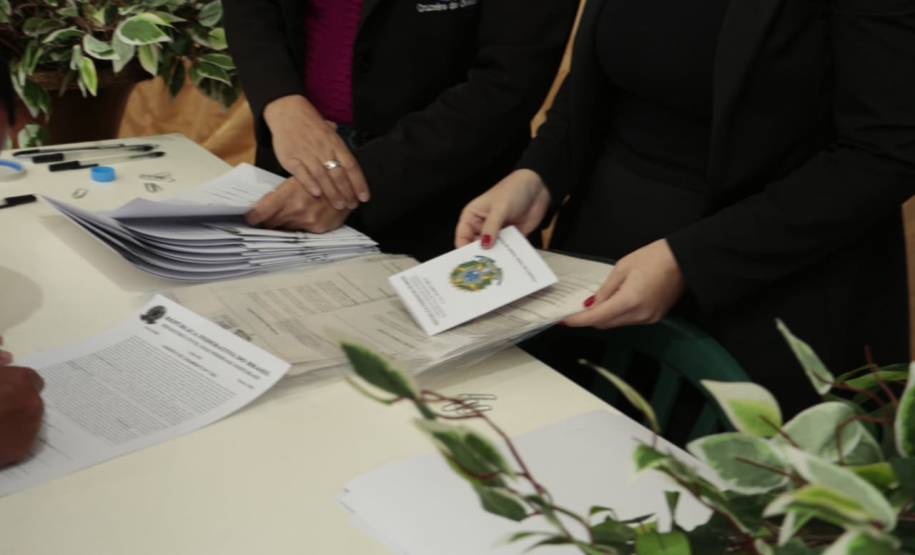 Detalhe de uma mesa com vários documentos impressos. Duas mulheres de terno preto manuseiam os papéis, e uma delas segura um livreto com o brasão da República do Brasil na capa. À esquerda, vê-se a mão de uma pessoa assinando um papel.