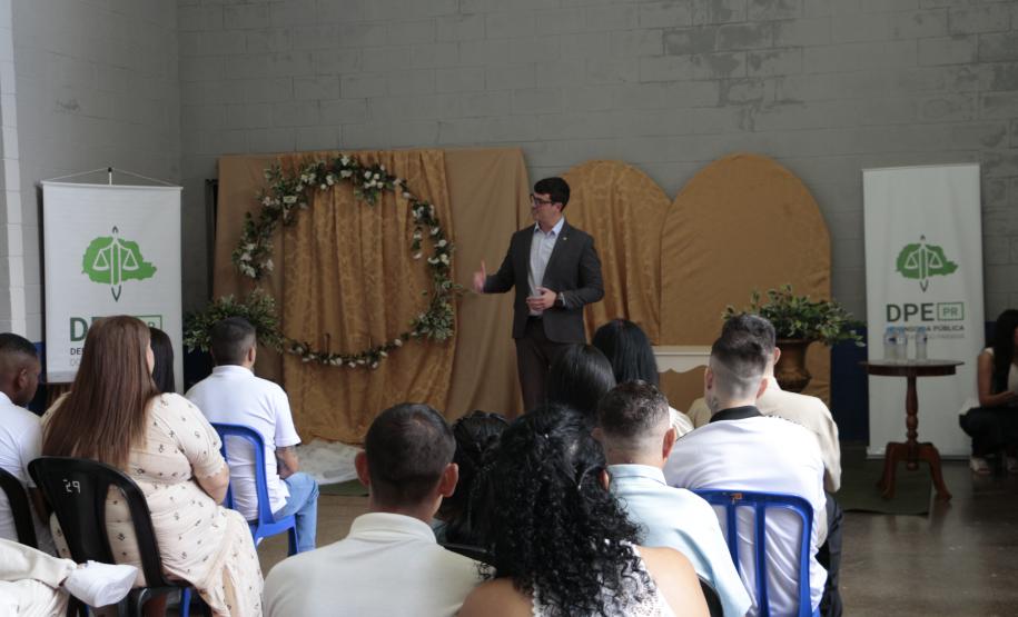 Um homem de terno escuro discursa em pé para o público acomodado em cadeiras de plástico azul e preta. Ele está em frente à decoração dourada com um arco de flores, ladeado por um banner da DPE-PR à direita.