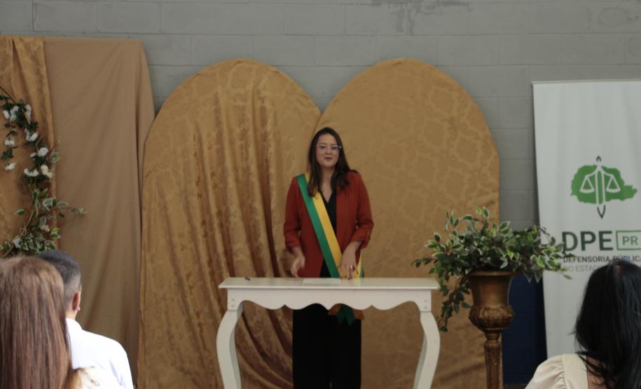 Uma mulher com blazer vermelho e a faixa transversal verde e amarela está em pé atrás da mesa branca, falando aos presentes. O cenário mantém os painéis dourados ao fundo e o banner da DPE-PR à direita.