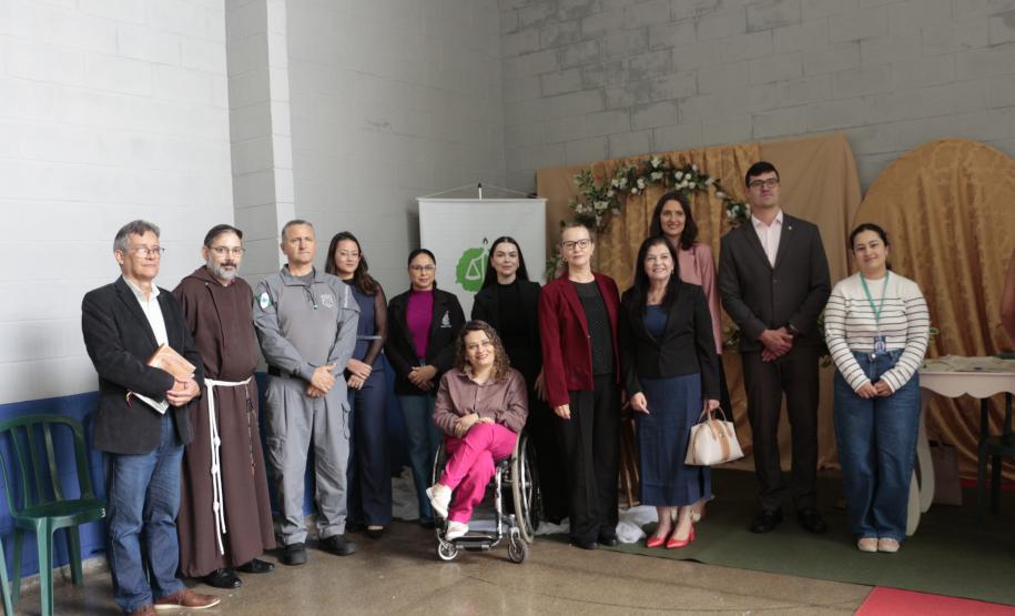 Foto posada de um grupo diversificado de doze pessoas em um espaço amplo. Estão presentes o policial penal, um frade de hábito marrom, uma mulher em cadeira de rodas e outras pessoas em trajes sociais, todos diante do painel dourado.