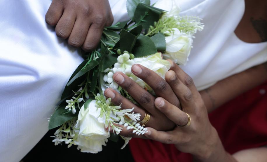 Detalhe em close de mãos negras entrelaçadas sobre um buquê de rosas brancas, ambas exibindo alianças douradas. Uma das pessoas veste um traje branco (vestido), e a outra usa uma blusa vermelha.