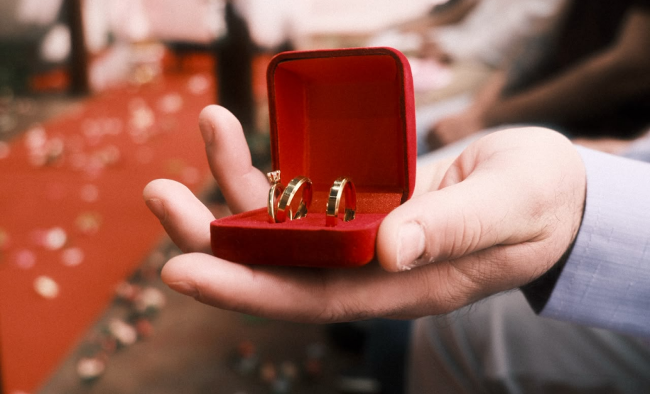 A mão de um homem, segura uma caixinha vermelha com três alianças de casamento.