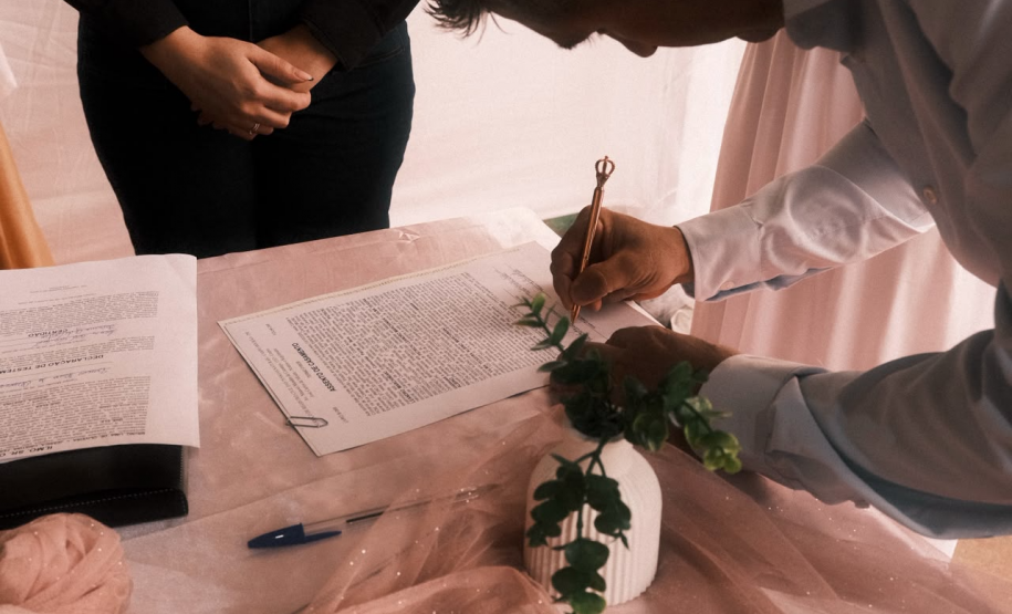 Homem, assinando documento em uma mesa de toalha branca. Ele está usando um terno e perto de si tem um pequeno vaso com uma rosa