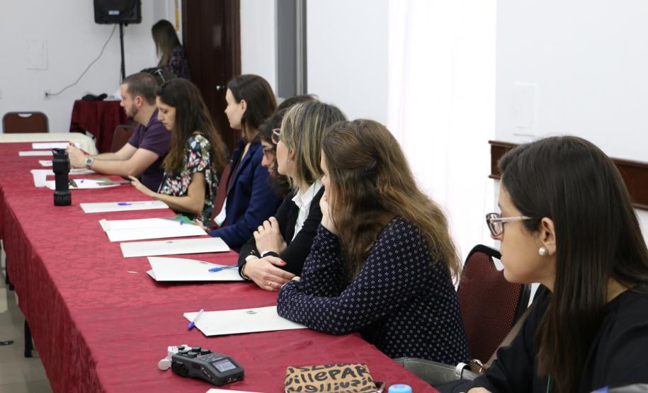 Na imagem vemos os participantes do II Encontro das Equipes Técnicas da DPPR sentados em uma mesa lateral comprida. Todos estão com um tanto virados para a direita, prestando atenção em quem fala.