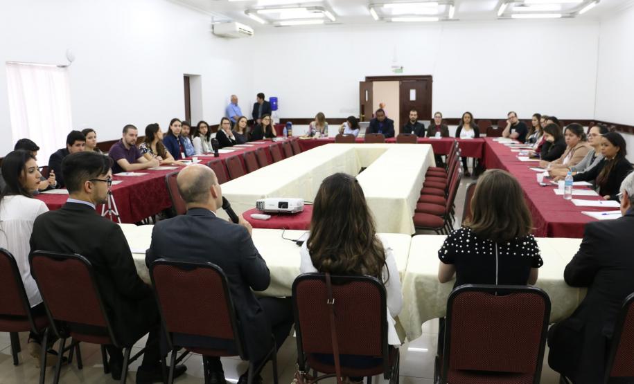 Vemos todos os participantes do encontro. A foto foi tirada atrás da mesa principal dos palestrantes, então vemos esses de costas e os participantes sentados em largas mesas laterais e uma frontal.