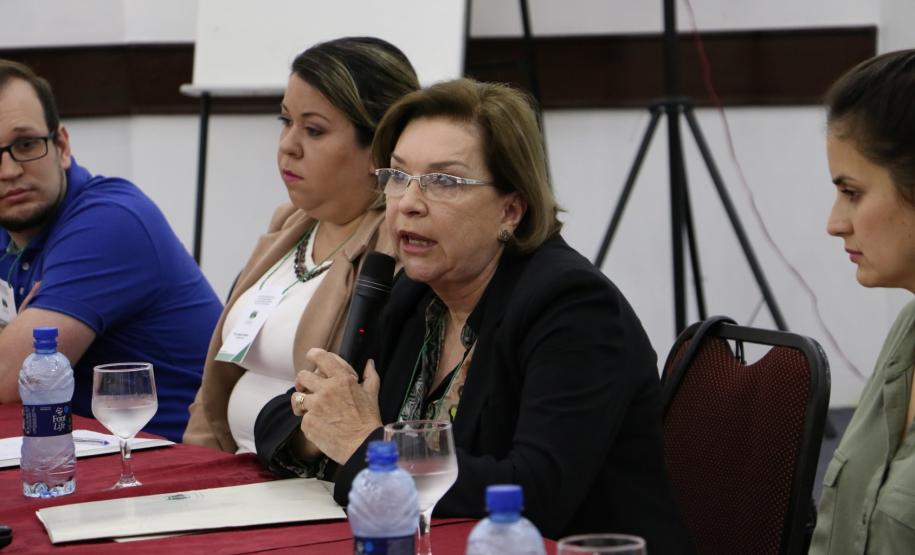 Maria Isabel, Assistente Social do CAM Curitiba, sentada na mesa de palestrantes com os outros falantes em seu lado.
A imagem foca apenas na Assistente Social e essa se encontra segurando um microfone próximo a sua boca.