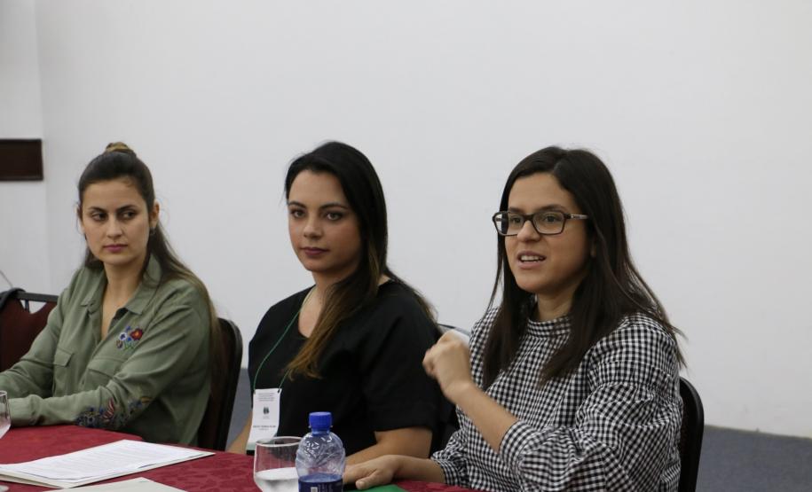 Temos as Assistentes Sociais do CAM Ponta Grossa, Ana Letícia de França e Evelyn Soares,ao fundo, sentadas no lado direito da mesa de falantes e  Taísa de Oliveira ,Assistente Social do CAM Curitiba, na ponta esquerda da mesa.