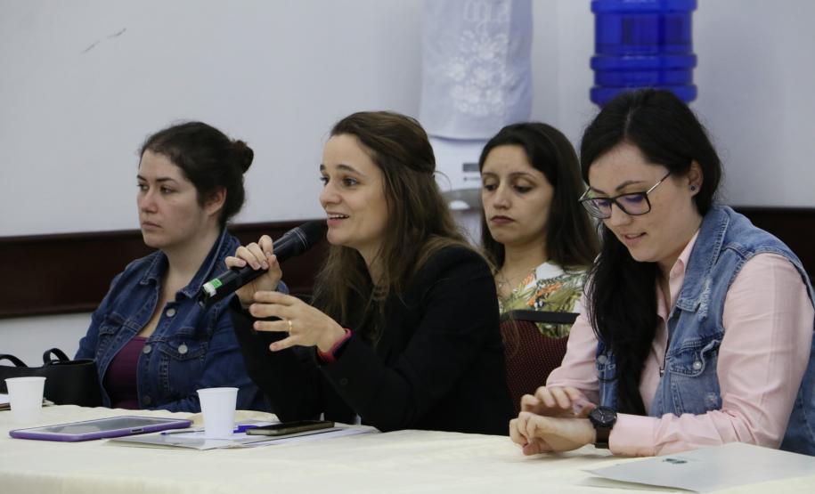 Vemos três participantes do evento. Camylla Basso, sentada centralmente, psicologa da Coordenação de Planejamento aparenta estar realizando uma pergunta.