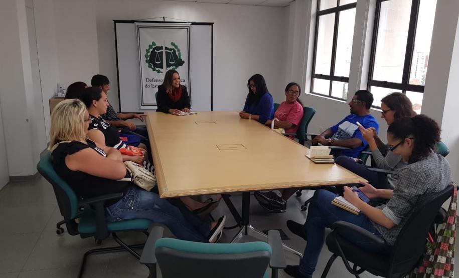 Mesa grande com representantes da Sociedade Barracão em volta e a dra. Olenka Lins ao fundo, do lado da bandeira da Defensoria Pública do Estado do Paraná.