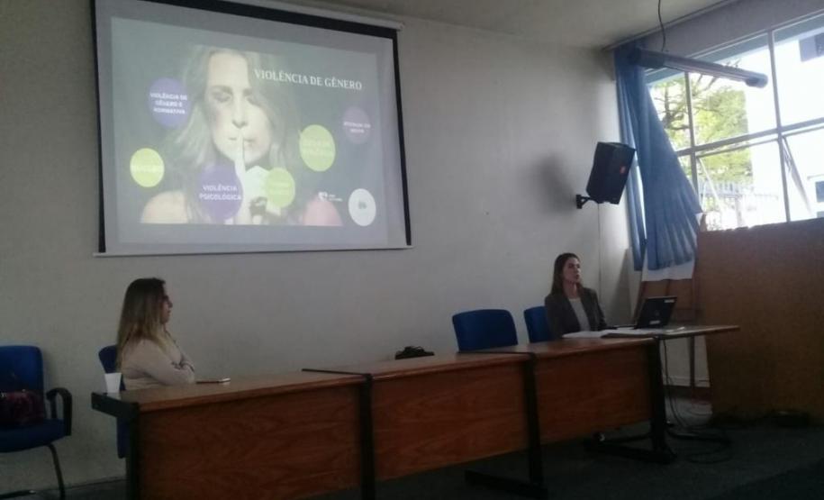A imagem consiste da Dra. Luciana Tramujas, coordenadora da defensoria da Casa da Mulher Brasileira, e da Dra. Eliana Lopes, Coordenadora do Pró-mulher, sentadas em uma mesa (cada uma em uma ponta) e ao fundo um tela com uma apresentação sobre violência e gênero.