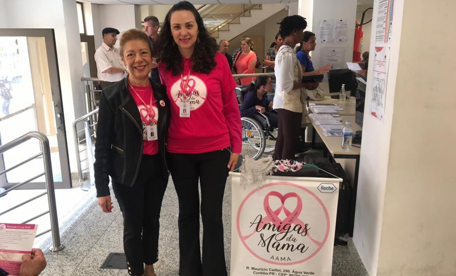 As duas palestrantes encontram-se juntas, no CAM Curitiba, após o termino da palestra. Ambas estão sorrindo para a foto e há um cartaz da Associação Amigas da Mama ao seu lado.