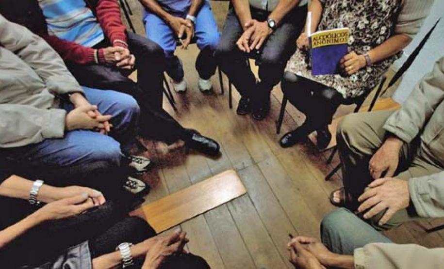 A foto foi tirada sob uma perspectiva de cima; Vemos um grupo de pessoas sentadas em roda, todas muito juntas, simulando uma reunião de AA.
