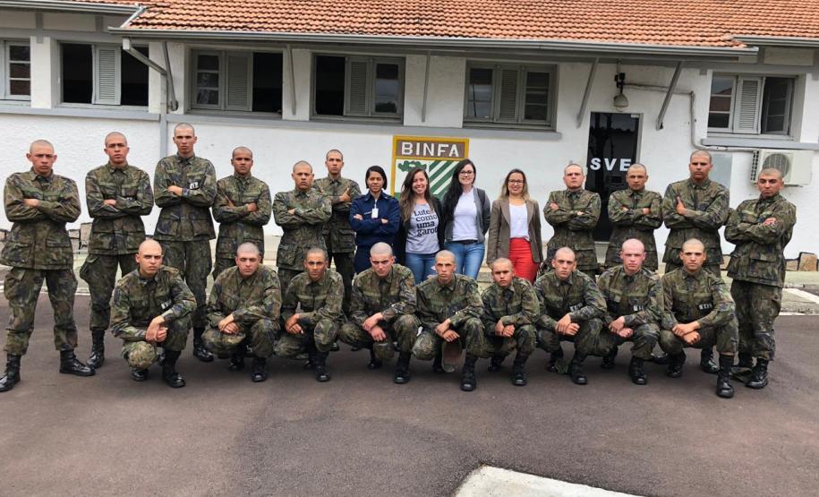 Vemos os soldados, alunos do curso, em fileiras - alguns abaixados e outros de pé - posando para foto. A equipe da DPPR da Casa da Mulher Brasileira se encontra em pé, no meio dos soldados,