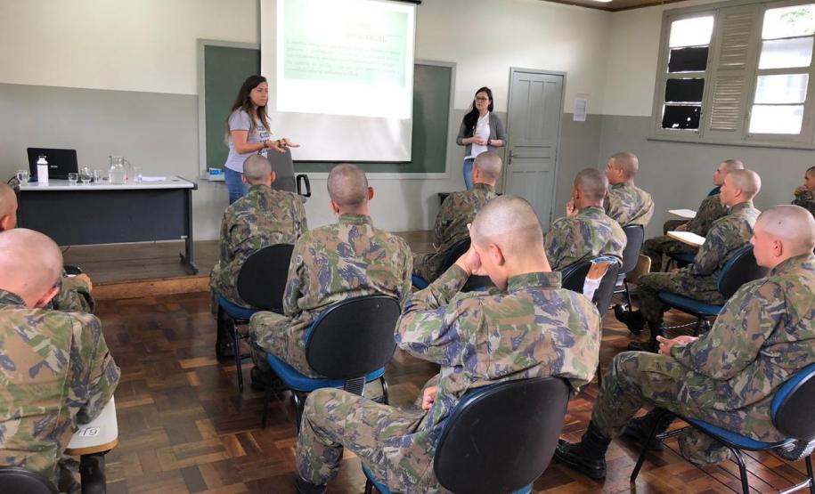 A foto foi tirada no fundo da sala. Nela vemos os soldados sentados em algumas certeiras prestando atenção na equipe da DPPR que está na parte da frente da sala, palestrando,