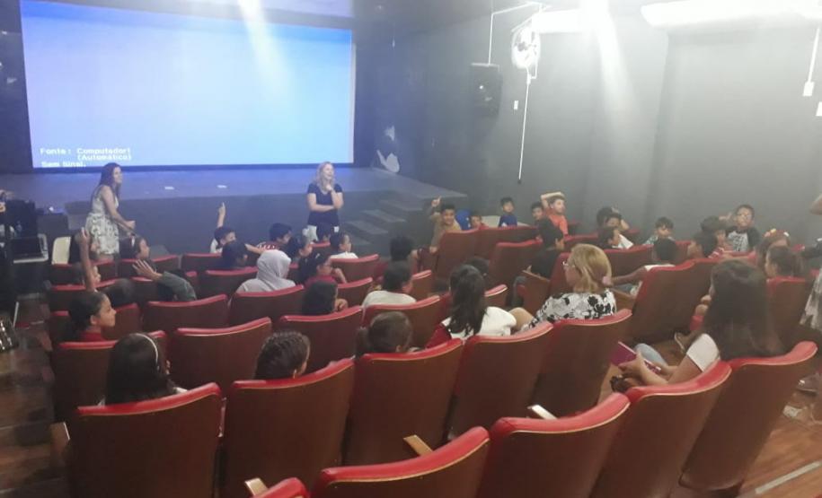 Na imagem vemos o encerramento do projeto no auditório da UNICENTRO. Temos os alunos sentados nas poltronas e as professoras em frente ao palco e telão.