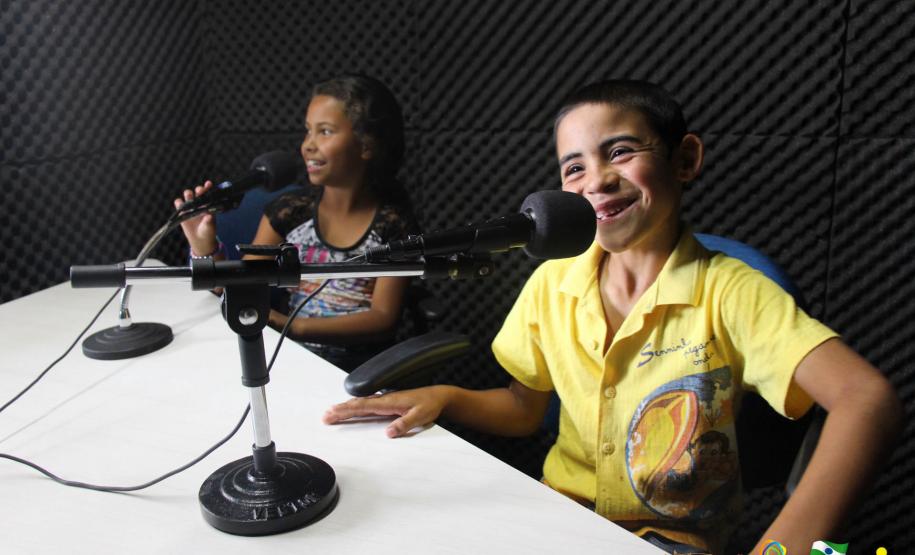 Na imagem vemos duas crianças em um estúdio de rádio, sentadas na mesa de gravação.