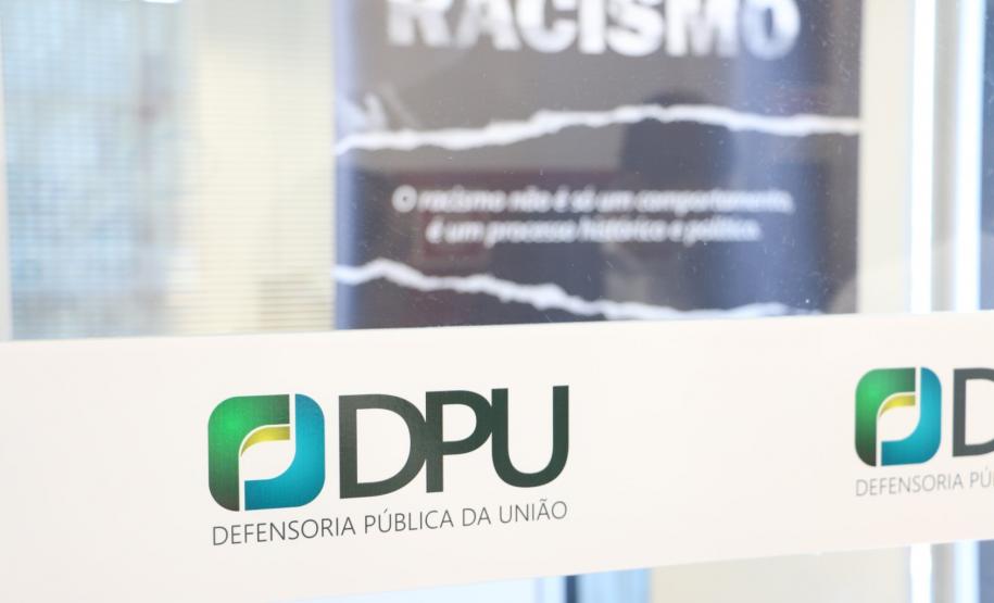 A foto foi tirada de uma porta transparente. onde está colada a logo da DPU. Essa se encontra em destaque na imagem.