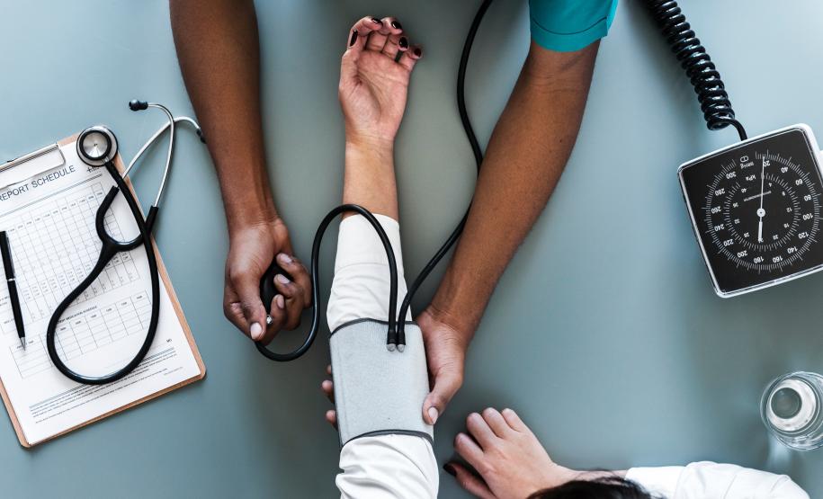 Imagem de um médico, onde esse está medindo a pressão do paciente.
 A foto encontra-se em uma perceptiva observada de cima, aparecendo apenas os braços de ambos.