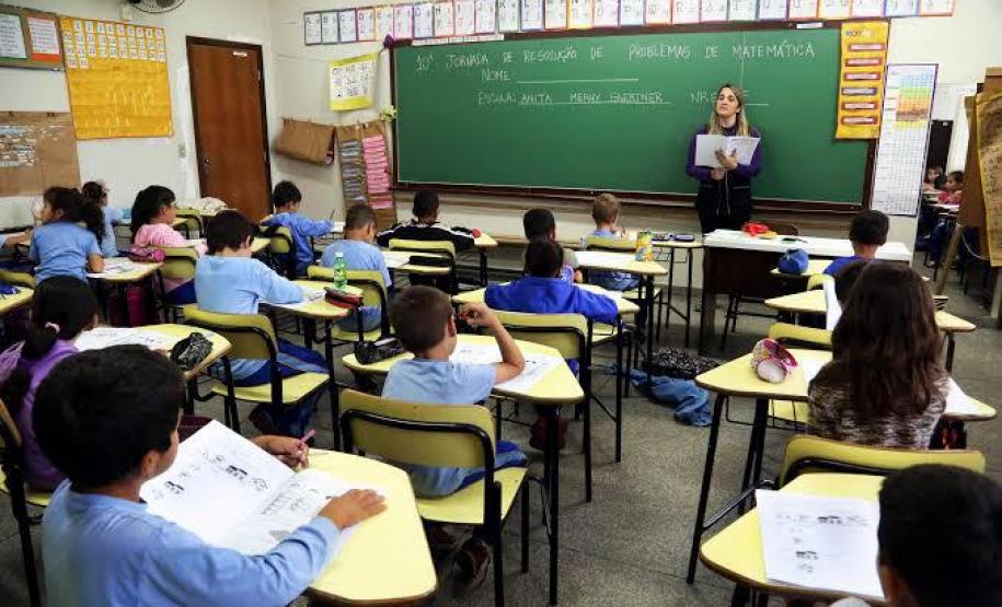 #LegendaAcessível: A imagem mostra a professora, na frente do quadro-negro ensinando para uma sala cheia de alunos, que acompanham a matéria pela apostila.