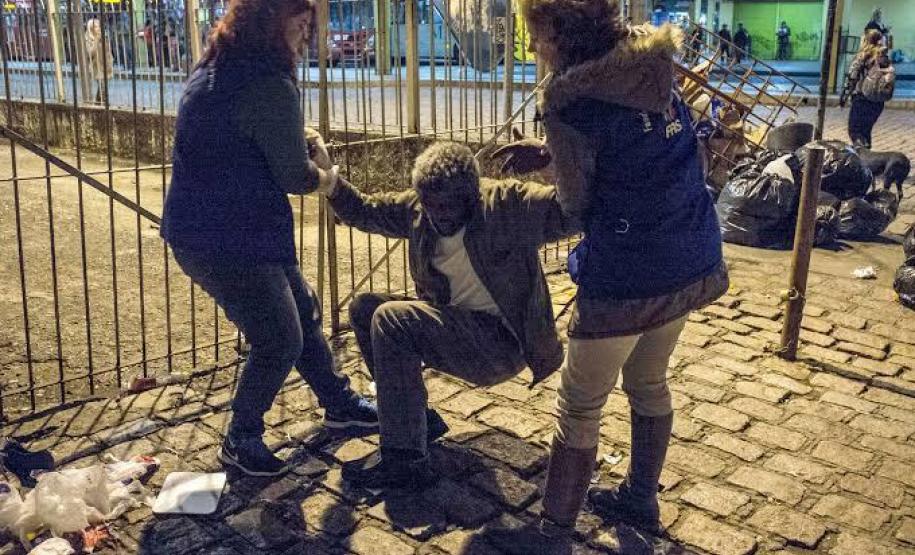 A imagem, uma foto ilustrativa, registra um morador de rua, idoso, sendo atendido, à noite, por duas assistentes sociais.